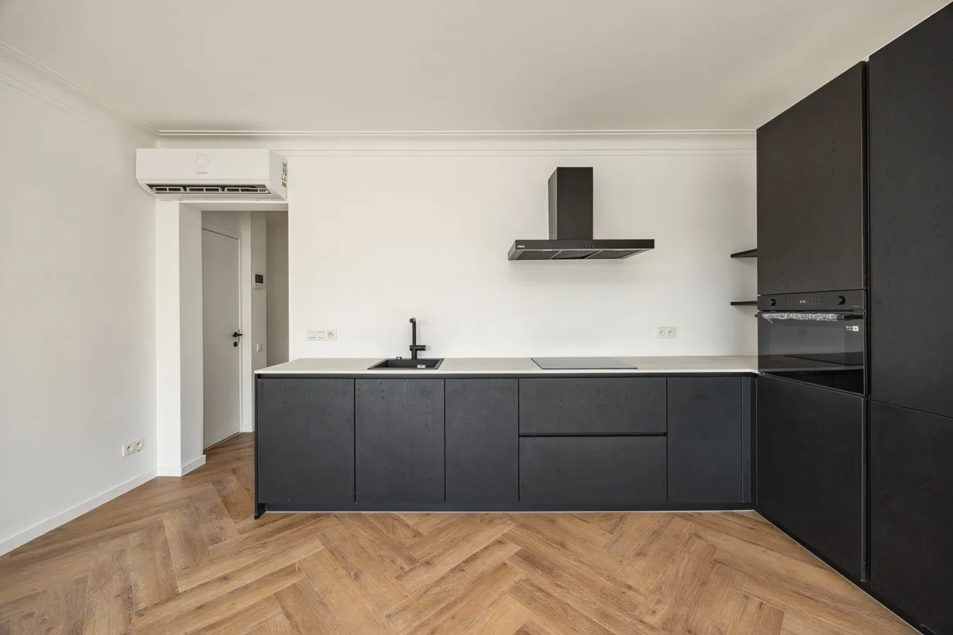 Strakke keuken met zwarte houten kasten, dampkap en inbouwoven op visgraatparket, met warmtepomp aan de muur, in Residentie Den Berm in Mortsel