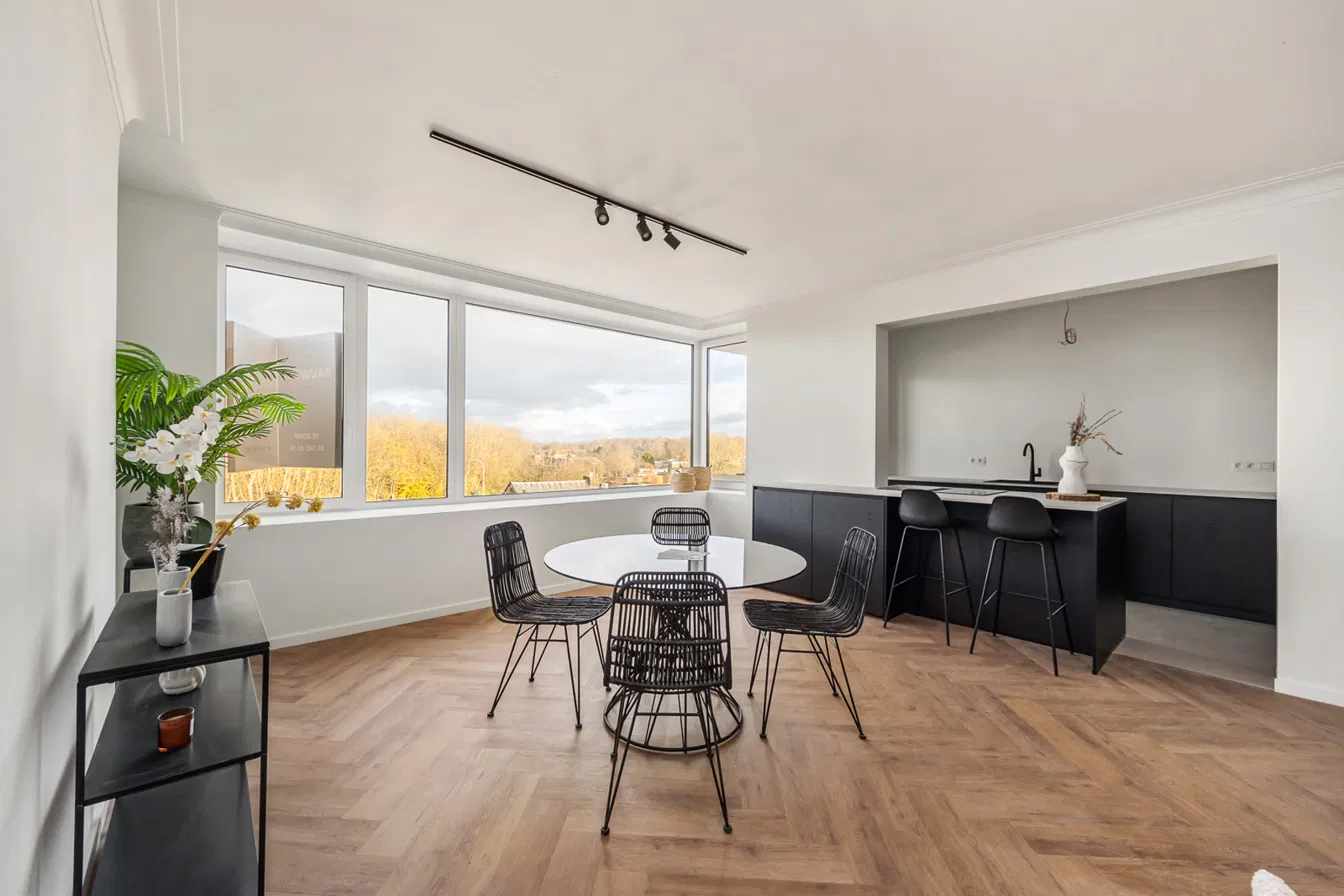Lichte leefruimte met ronde eettafel, visgraatparket en zwarte keuken met bar, met groot hoekraam en zicht op het groen in Residentie Den Berm in Mortsel