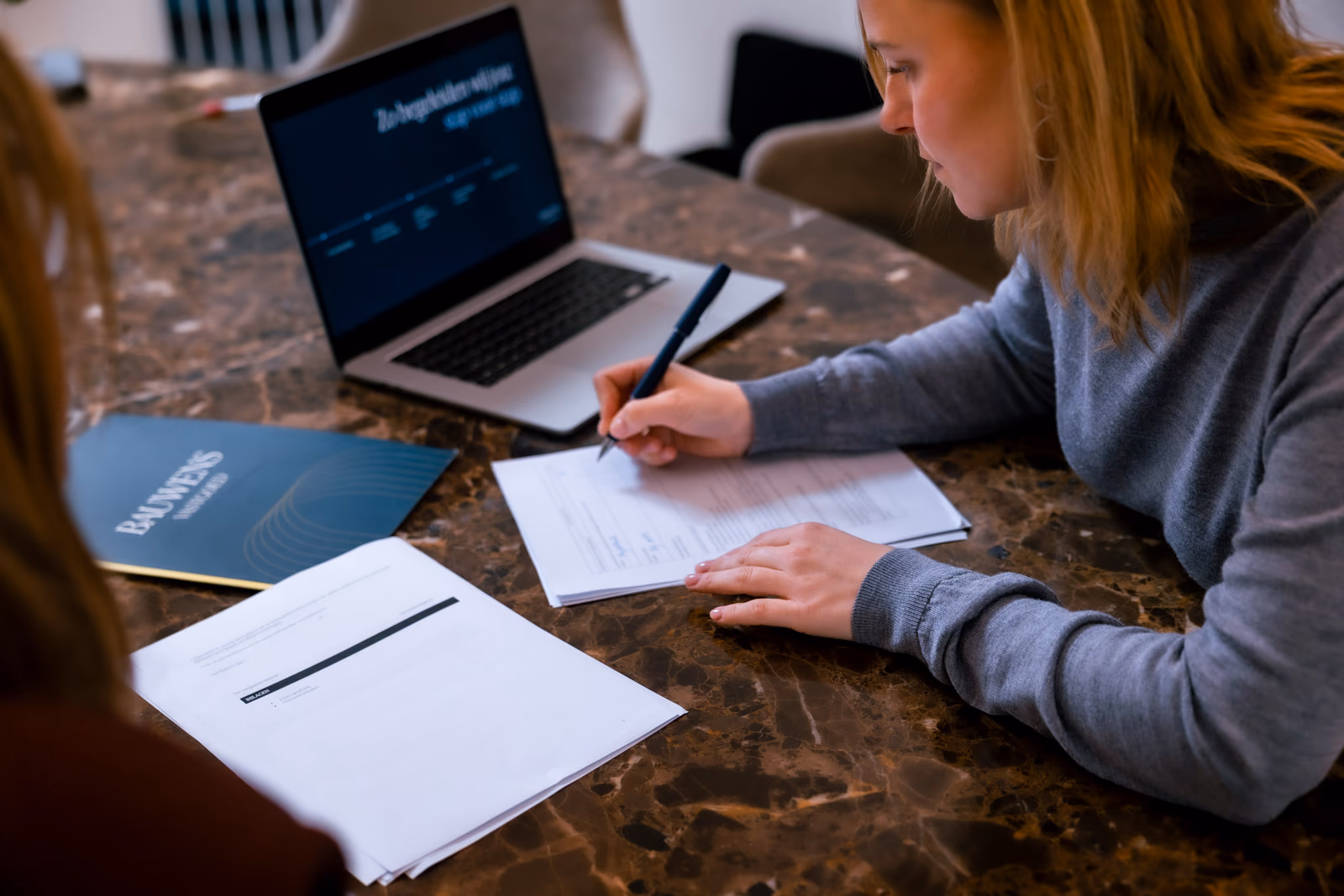 Vrouw vult aan een marmeren tafel een document in naast een laptop en een map van Bauwens Vastgoed.