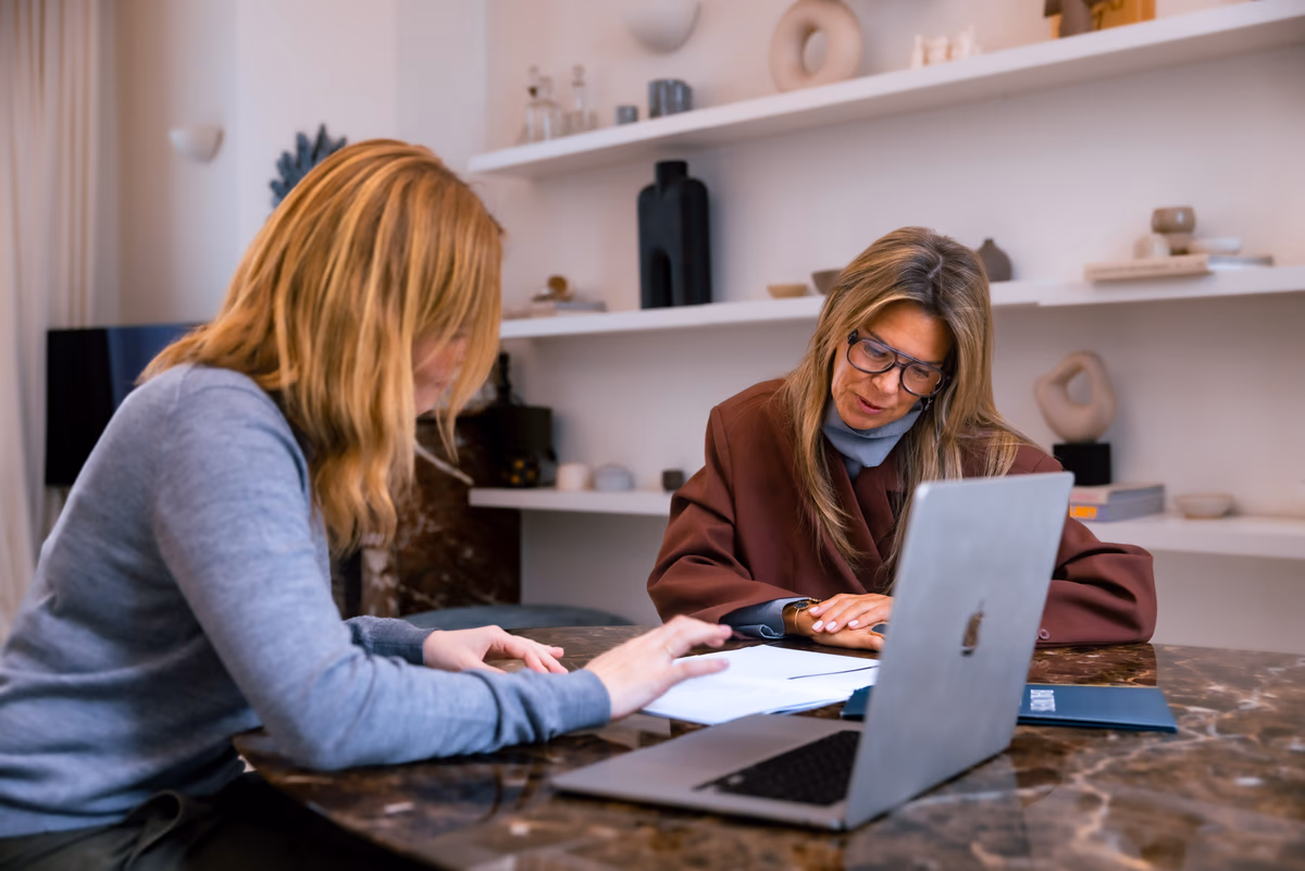 Vrouw vult aan een marmeren tafel met een andere vrouw een document in naast een laptop en een map van Bauwens Vastgoed.